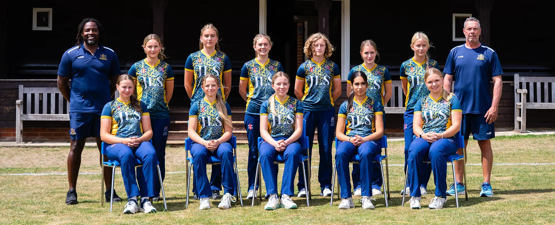 A smartly dressed senior girls’ cricket team, with coaches standing either side, posing in front of the pavilion.